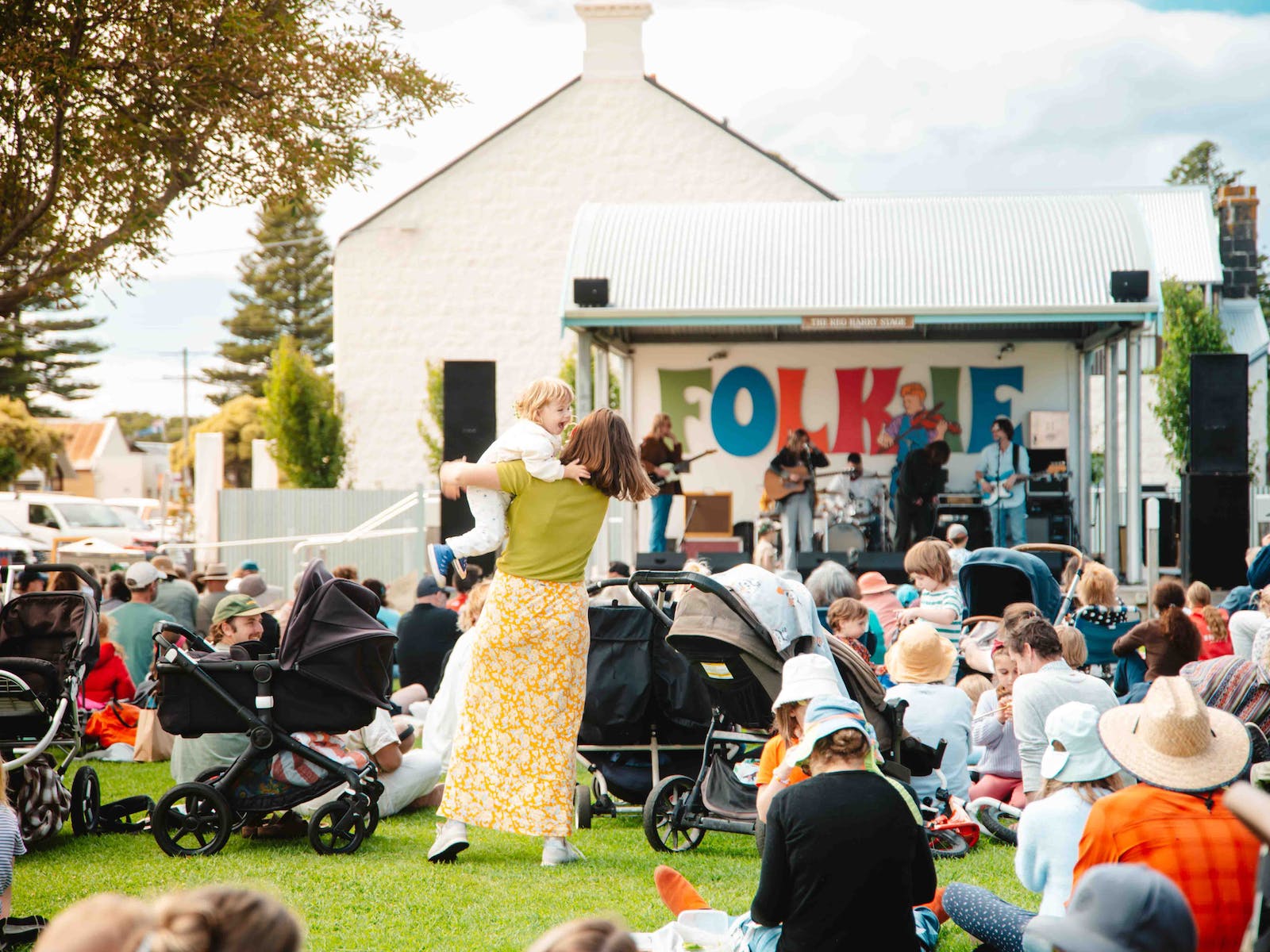 A child and a woman dance together outside watching a band. There are many other people around.